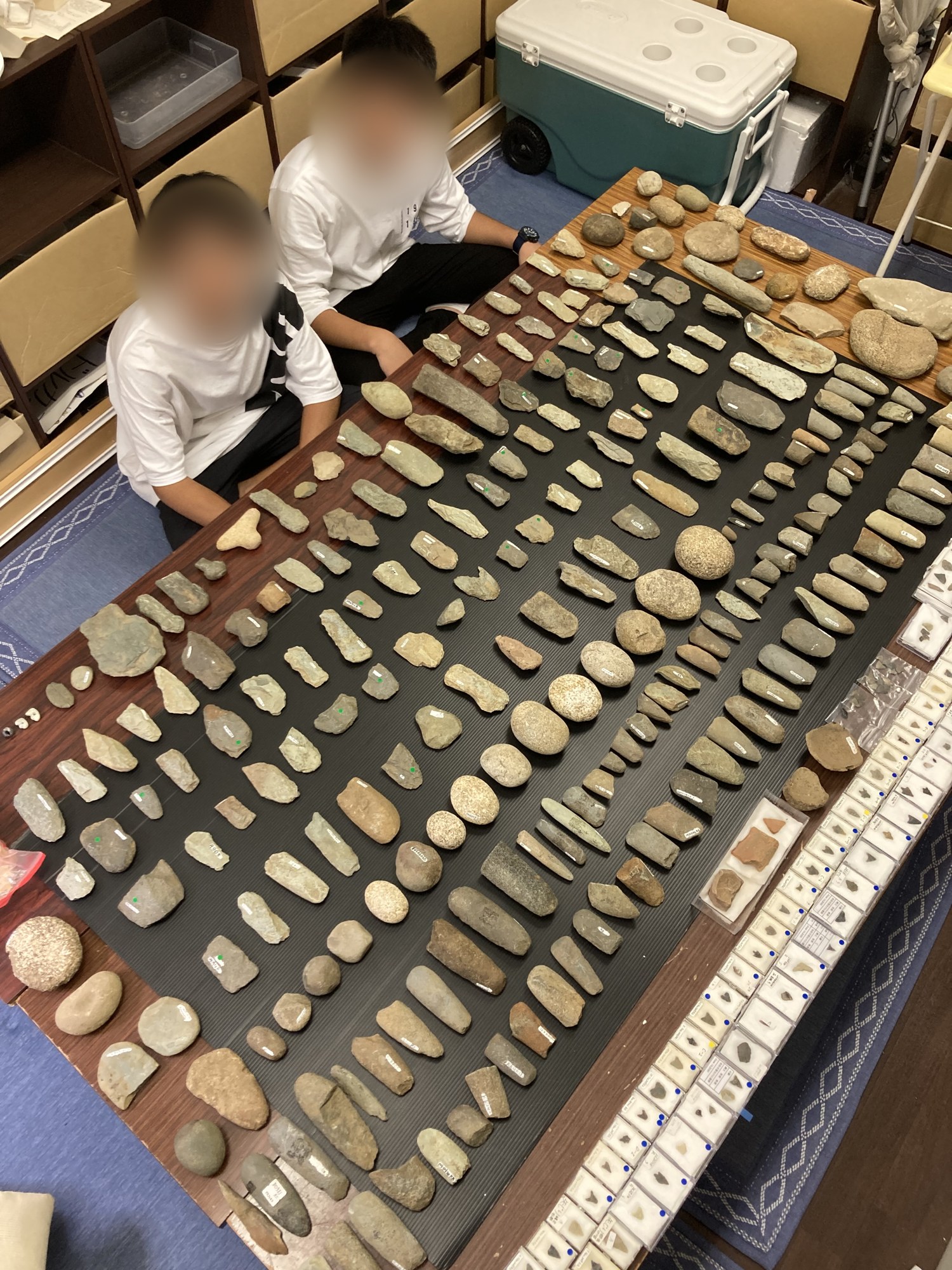 机いっぱいに並べた石器と、その横に座る2人（顔はぼかし加工）